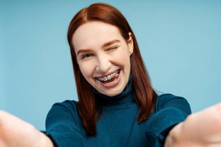 Photo for Closeup selfie of a joyful redhead woman with braces, wearing a polo neck sweater, capturing herself and looking at the camera, isolated on a blue background - Royalty Free Image