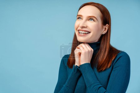 Photo for Happy ginger haired woman with braces, wearing a polo neck sweater, joins her hands in supplication, looking away, isolated against blue background with copy space - Royalty Free Image