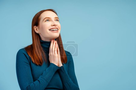 Photo for Smiling beautiful red haired in a polo sweater, braces visible, clasps hands in a prayerful gesture, looking away, isolated on a blue background - Royalty Free Image