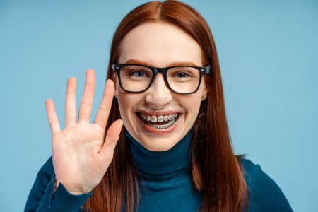 A cheerful ginger haired young woman in glasses and braces, clad in a polo neck sweater, waves hello, her smile lighting up the photo, isolated on a blue background