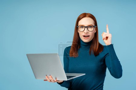 Photo for Shot of an attractive redhead with braces and eyeglasses, holding a laptop and showing a light bulb moment with her index finger, surprised expression, isolated on blue background - Royalty Free Image