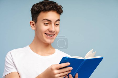 Smart smiling school boy wearing eyeglasses holding book reading isolated on blue background. College student studying, learning language. Education concept 