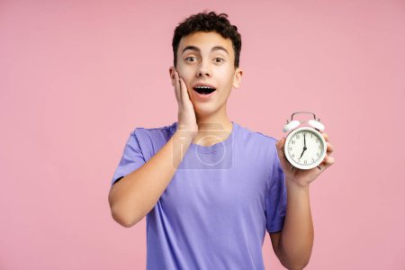 Shocked boy with dental braces holding alarm clock and screaming isolated against pink background. Emotional student late to study 