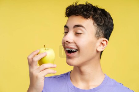 Curly smiling boy with braces in purple shirt holding green apple isolated on yellow background studio portrait. Healthcare procedures concept 