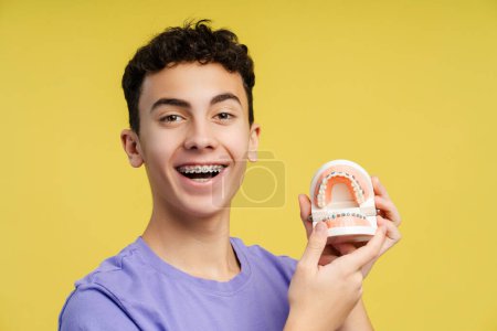 Close up shot of a smiling, curly haired teenager with braces, holding dental molds, laughing, isolated on yellow background. Emphasizes orthodontic therapy concept 