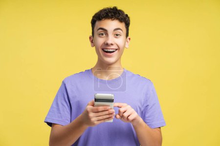 Photo for Portrait of amazed teenager wearing braces holding mobile phone, using website, looking at camera isolated on yellow background. Technology concept - Royalty Free Image