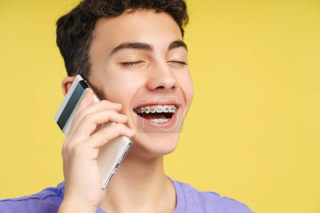 Closeup portrait of smiling boy with braces talking on mobile phone with closed eyes isolated on yellow background. Happy modern hipster answering call. Technology concept 