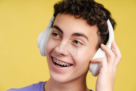 Foto de Retrato de un adolescente sonriente y atractivo con frenos dentales, escuchando música en auriculares inalámbricos aislados sobre fondo amarillo - Imagen libre de derechos