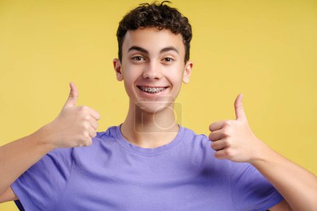 Téléchargez les photos : Portrait d'un beau garçon souriant, adolescent de 15 ans avec des accolades dentaires portant un t-shirt élégant décontracté violet montrant les pouces vers le haut en regardant la caméra isolée sur fond jaune. Concept de publicité - en image libre de droit