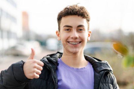 Portrait of attractive boy, teenager wearing stylish casual clothes showing thumb up, cool gesture looking at camera standing on urban street. Advertisement concept 