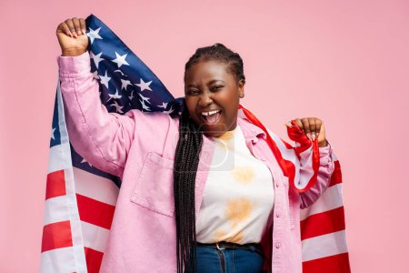 Foto de Retrato muy alegre mujer afroamericana sosteniendo la bandera de EE.UU. aislado sobre fondo rosa. Estudiante feliz mirando a la cámara, la educación en EE.UU.. Concepto de inmigración - Imagen libre de derechos