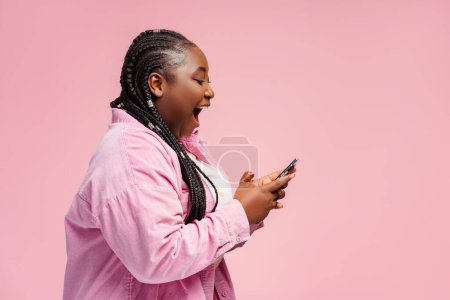 Foto de Mujer africana feliz celebración de teléfonos inteligentes utilizando la aplicación móvil, compras en línea con la venta, dinero en efectivo aislado en el fondo rosa, espacio de copia. Apuestas deportivas femeninas muy alegres, ganar dinero, leer buenas noticias - Imagen libre de derechos