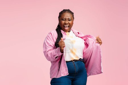 Téléchargez les photos : Portrait ravi femme afro-américaine avec des tresses élégantes portant chemise rose à la mode isolé sur fond. Heureux Nigérian plus la taille modèle danse célébration succès. Concept positif du corps - en image libre de droit