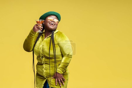 Foto de Retrato de mujer afroamericana con estilo feliz sosteniendo vidrio con cóctel mirando hacia otro lado aislado sobre fondo amarillo, espacio de copia. Sonriente mujer bebiendo alcohol. Concepto de fiesta de cumpleaños - Imagen libre de derechos