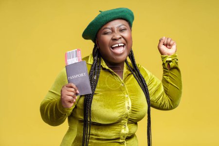 Portrait overjoyed stylish African woman holding passport with frights ticket isolated on background. Happy tourist waiting for travel. Vacation, advertisement concept