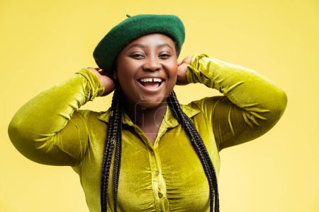 Photo for Beautiful smiling plus size fashion model with stylish braids hair, wearing green beret isolated on yellow background. Portrait happy African woman posing in trendy outfit in studio. Shopping, sale - Royalty Free Image