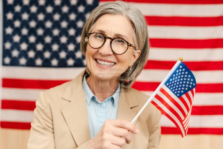 Portrait of smiling confident senior woman, politician holding American flag looking at camera. Vote, United States presidential election concept 