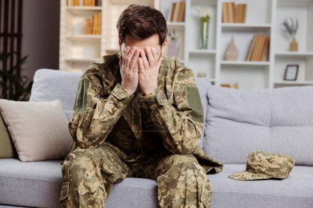 Man in army clothing, home but facing depression. He is sitting on the living room sofa, covering his face with his hands. Focus on mental support for veterans