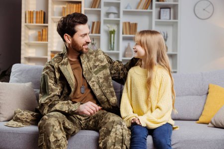 Téléchargez les photos : Bonne famille militaire à la maison. Père souriant en uniforme de l'armée touchant doucement la joue de sa petite fille. Ils sont assis sur le canapé, se regardant dans les yeux. Célébration du concept de retour des soldats - en image libre de droit