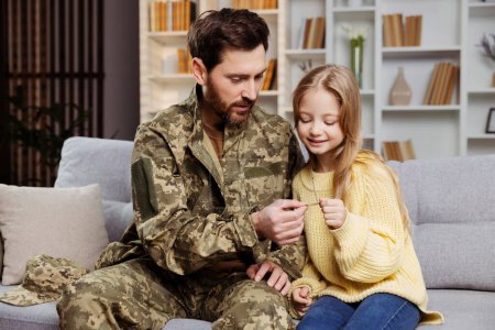 Téléchargez les photos : Bonne famille militaire à la maison. Père joyeux en camouflage, présentant son badge à sa petite fille. Assis sur le canapé ensemble. Concept de retour des soldats - en image libre de droit