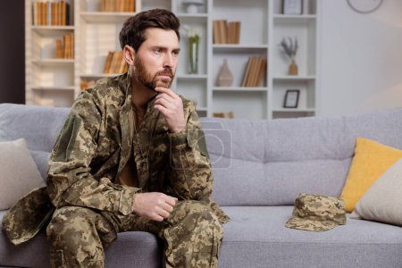 Photo for Man in military uniform, returning home, battling depression. He is sitting on a couch and looking away with melancholy. Focus on mental support for veterans - Royalty Free Image
