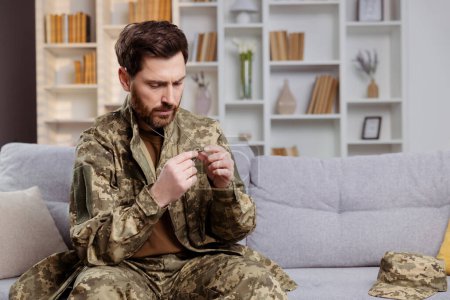 Soldier in military uniform, back home with a heavy heart. Sitting on the couch, he looking at his dog tag, thinking. Theme of psychological help for servicemen