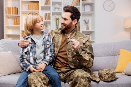 Feliz familia militar en el interior, con el padre sonriente en uniforme, sosteniendo a su orgulloso hijo y mostrando su identificación. Están sentados en un sofá, abrazando