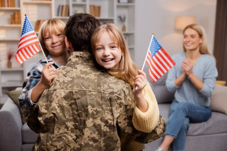 Téléchargez les photos : Heureuse famille américaine célébrant le retour d'un soldat. Père en tenue militaire, embrassant ses joyeux enfants, qui le saluent avec des drapeaux américains. Dans leur salon, mère excitée attend sur le canapé - en image libre de droit