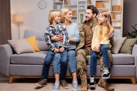 Photo for Happy military family at home. Smiling father in uniform, beautiful mother, and little children sitting on the sofa together, looking at each other. Concept of soldier homecoming - Royalty Free Image