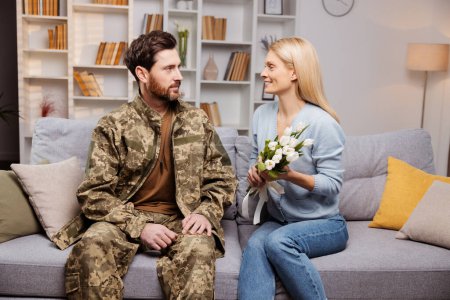 Téléchargez les photos : De retour du service. Mari souriant en tenue militaire regardant avec amour sa femme joyeuse. Assise sur le canapé, elle tient des fleurs, toutes les deux heureuses à la maison ensemble - en image libre de droit