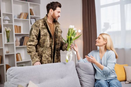 Photo for Smiling serviceman in army clothes returning home and giving flowers to excited woman, sitting on a sofa. Soldier homecoming concept - Royalty Free Image