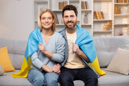Téléchargez les photos : Heureuse famille ukrainienne dans leur maison, arborant fièrement le drapeau ukrainien, les mains entrelacées. Couple marié souriant sur le canapé du salon. Adopter le concept d'Ukraine - en image libre de droit