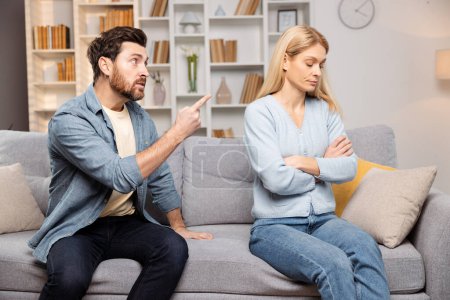 Photo for Living room family dispute: an angry man shouting at his wife, sitting with arms crossed on the sofa, indifferent to his rage - Royalty Free Image
