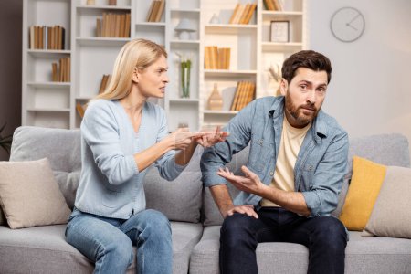 Photo for Annoyed wife counting off problems on her fingers at her husband, sitting on sofa in their living room. Man looking at camera, demonstrating his misunderstanding. Captures a moment of marital discord - Royalty Free Image