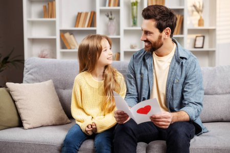 Photo for Happy family celebrating Father's Day. Smiling father holding a handmade card from his little daughter, both sitting on the living room sofa, looking at each other - Royalty Free Image