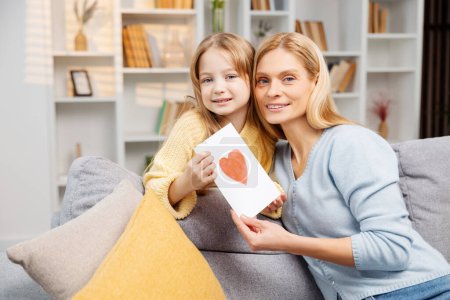 Photo for Happy family celebrating Mother's Day. A lovely mother and her young daughter with a handmade card looking at the camera, enjoying the moment on their sofa - Royalty Free Image