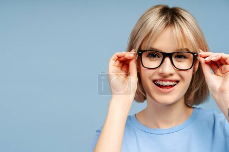 Téléchargez les photos : Portrait d'une femme blonde confiante avec des bretelles, portant des lunettes souriantes et regardant la caméra, sur un fond bleu - en image libre de droit