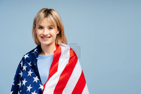 Téléchargez les photos : Souriant belle jeune femme aux cheveux blonds enveloppé dans le drapeau américain en regardant la caméra, l'espace de copie. Femme supporter debout isolé sur fond bleu. Concept de fête de l'indépendance américaine - en image libre de droit