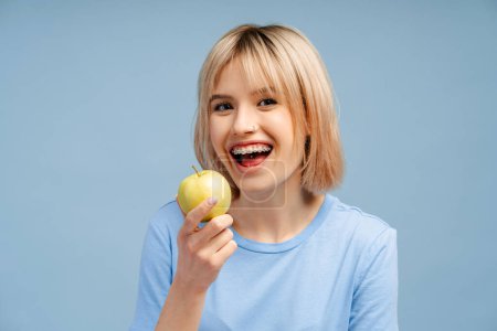 Photo for Closeup shot of gorgeous blonde haired female wearing braces, holding an apple, isolated on a blue background. Highlights oral care - Royalty Free Image
