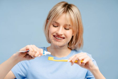 Téléchargez les photos : Belle femme avec des bretelles tenant versant du dentifrice sur une brosse à dents isolée sur fond bleu. Concept quotidien de routine et d'hygiène matinale - en image libre de droit