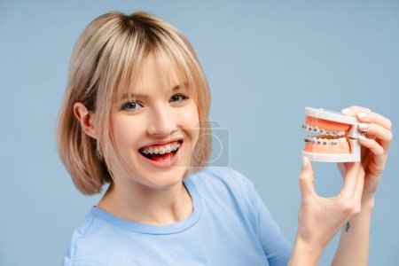 Téléchargez les photos : Joyeux jeune femme mignonne avec des appareils dentaires sur les dents tenant modèle en plastique mâchoire humaine avec des appareils dentaires isolés sur fond bleu. Dentisterie, soins de santé, concept de traitement orthodontique - en image libre de droit