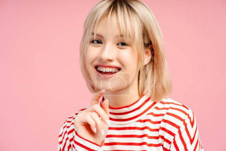 Photo for Attractive happy young woman with blond hair, with braces, smiling, looking at camera. Cheerful female student isolated on pink background. Concept of dental care, advertisement - Royalty Free Image