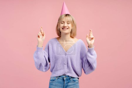Foto de Mujer atractiva feliz, con gorra de fiesta cruzando los dedos mientras espera el presente aislado sobre fondo rosa. Concepto de cumpleaños de fiesta y celebración - Imagen libre de derechos