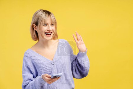 Photo for Smiling beautiful woman holding mobile phone greeting, isolated on yellow background. Happy blogger influencer waving hand. Technology, social media concept - Royalty Free Image