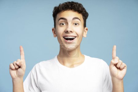 Photo for Confident, excited teenage boy with braces showing finger on copy space, hands up standing isolated on blue background. Concept of advertisement, shopping - Royalty Free Image