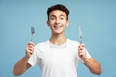Photo for Smiling attractive boy holding fork and knife looking at camera. Hungry teenager standing isolated on blue background. Nutrition concept - Royalty Free Image