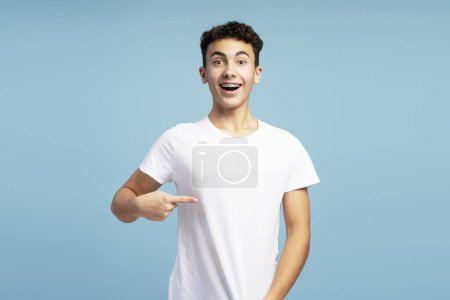 Téléchargez les photos : Un garçon souriant et excité portant un t-shirt blanc, pointant du doigt elle, se maquillant, regardant la caméra. Adolescent attrayant isolé sur fond bleu. Concept de publicité - en image libre de droit