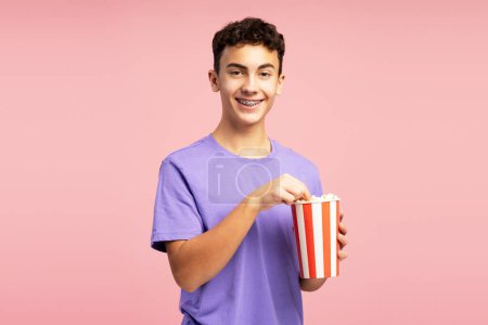 Photo for Smiling, handsome boy holding bucket of popcorn, eating, looking at camera. Excited teenager standing isolated on pink background. Concept of entertainment, snacks - Royalty Free Image