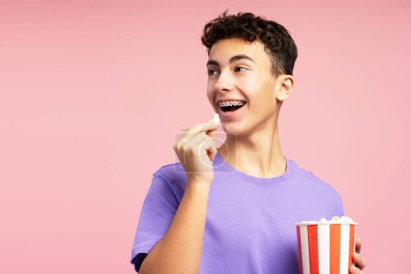 Téléchargez les photos : Adolescent excité et attrayant mangeant du pop-corn, tenant un seau regardant loin de l'espace de copie. Beau garçon avec des bretelles regardant un film. Concept de divertissement - en image libre de droit