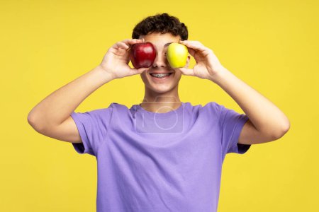 Foto de Un tipo guapo y divertido con aparatos ortopédicos sosteniendo manzanas aisladas sobre un fondo amarillo. Adolescente atractiva divirtiéndose. Concepto de comida saludable - Imagen libre de derechos